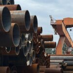 Stacked steel pipes beside an industrial crane in an outdoor yard, showcasing heavy industry and construction materials.